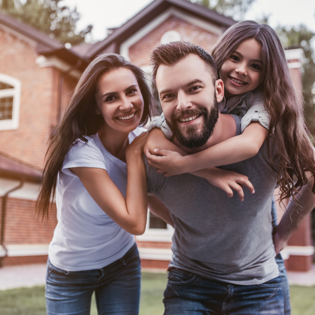 A joyful family poses in front of a brick house. A man carries a smiling girl on his back, while a woman stands beside them, all expressing happiness.